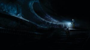 Two figures in bulky spacesuits approach a bio-mechanical, twisting structure that rises slowly up in front of them, they are shining flashlights on it.