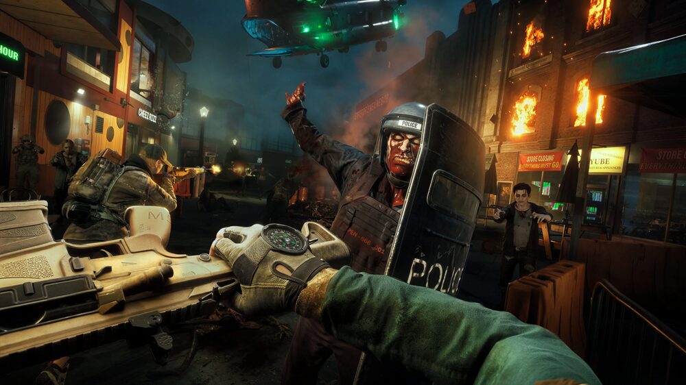A first-person view showing the PoV character about to hit a zombie in riot gear carrying a shield with the butt of their gun. In the background a modern city is on fire and other uninfected humans are fighting zombies.